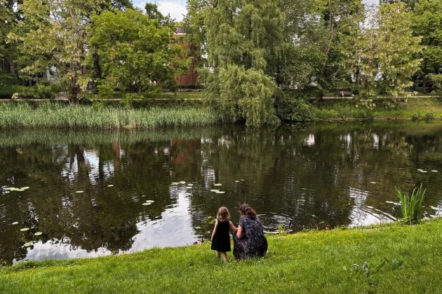 Kvinna och barn står på gräsmatta och tittar på en damm i parkmiljö.